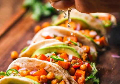 A hand squeezes lime juice over a row of veggie tacos on a wooden board, garnished with ingredients like chickpeas, avocado, and cilantro.