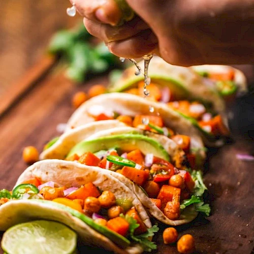 A hand squeezes lime juice over a row of veggie tacos on a wooden board, garnished with ingredients like chickpeas, avocado, and cilantro.