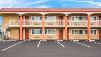 The image shows a two-story motel with exterior corridors, multiple red doors, and an empty parking lot in front of it.