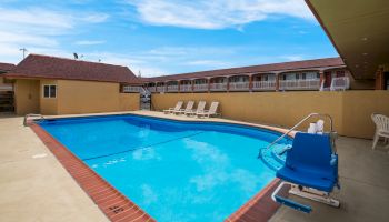 An outdoor swimming pool with lounge chairs, a pool lift, and a building in the background under a clear blue sky.