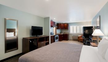 A hotel room with a bed, TV, mirror, and a kitchenette with wooden cabinets and appliances, plus a cozy seating area with a chair and lamp.