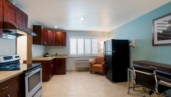 The image shows a modern kitchen and living area with wooden cabinets, a stove, fridge, chair, desk, and a window with blinds, ending the sentence.