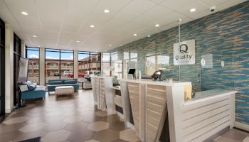 The image shows a modern hotel lobby with a reception desk, seating area, and large windows. The decor includes a blue accent wall and tiled floor.