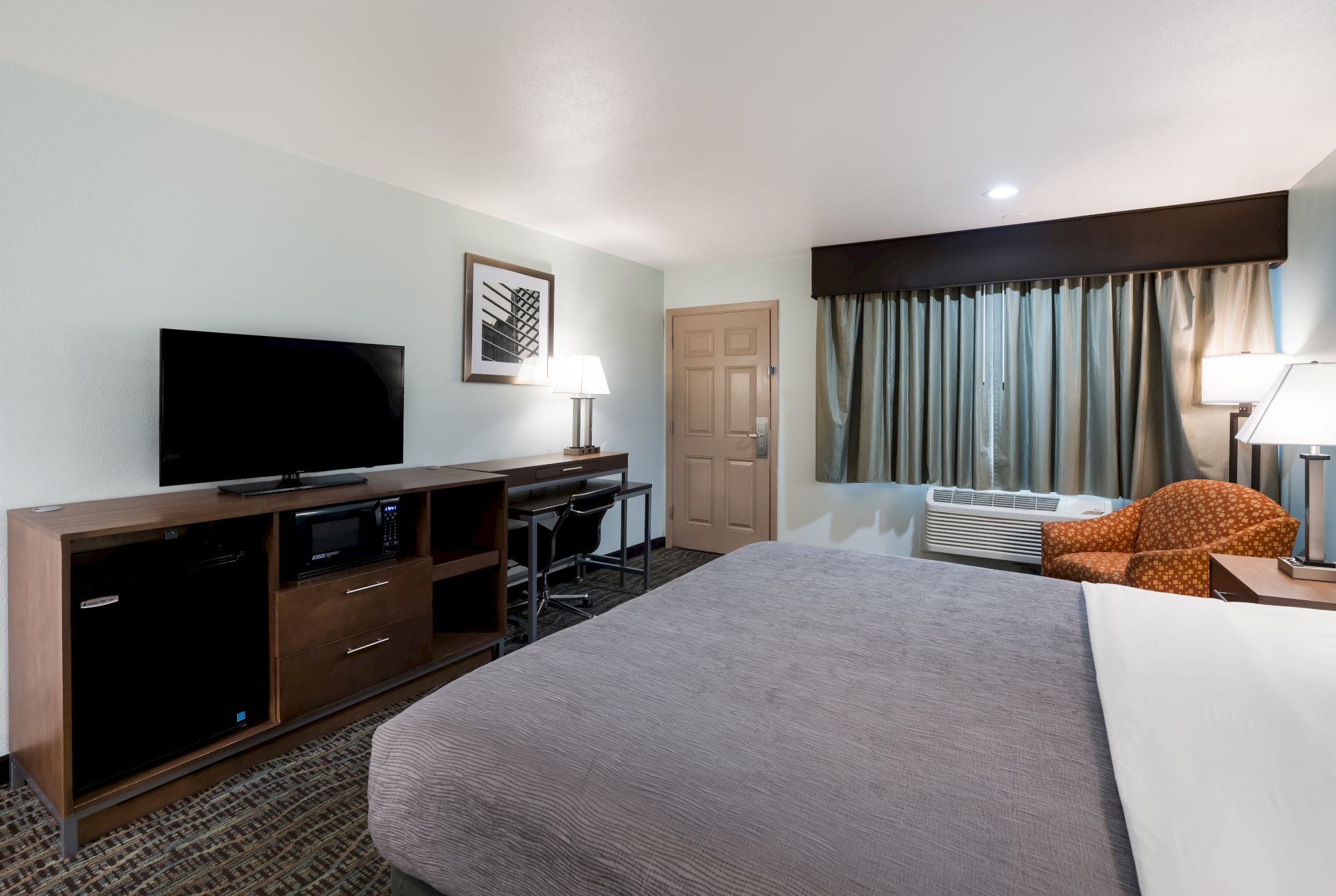 A hotel room with a bed, TV, desk, chair, and curtains. There is a lamp on the desk and another by the chair near the window.