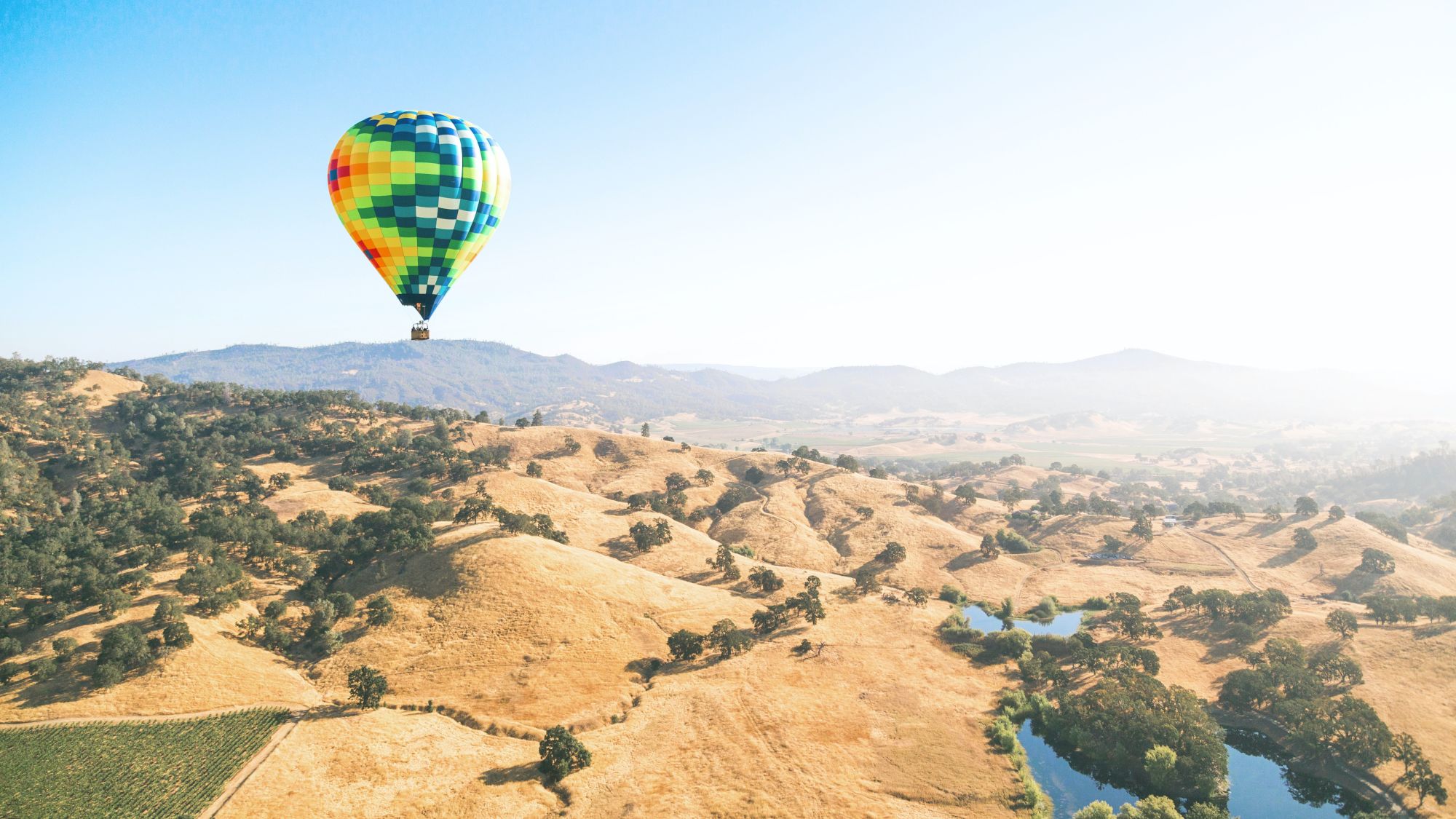 A colorful hot air balloon floats over a picturesque landscape with rolling hills, scattered trees, and a body of water on a clear, sunny day.