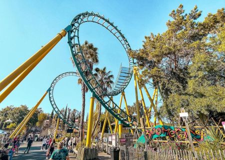 The image shows a vibrant amusement park with a roller coaster featuring loops and yellow supports, surrounded by trees and people walking around.