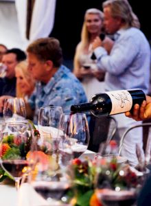 People are seated at a long table, enjoying food and wine, while one person pours wine into glasses and another speaks on a microphone.