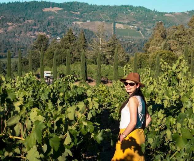 A person in a vineyard, wearing sunglasses and a hat, with hills and trees in the background on a sunny day.