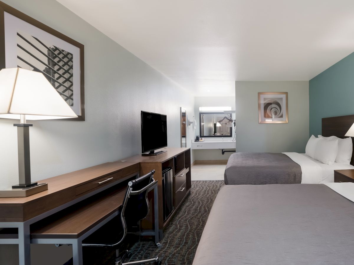 The image shows a hotel room with two beds, a desk with a lamp and chair, a TV on a dresser, and a mirror in the background.