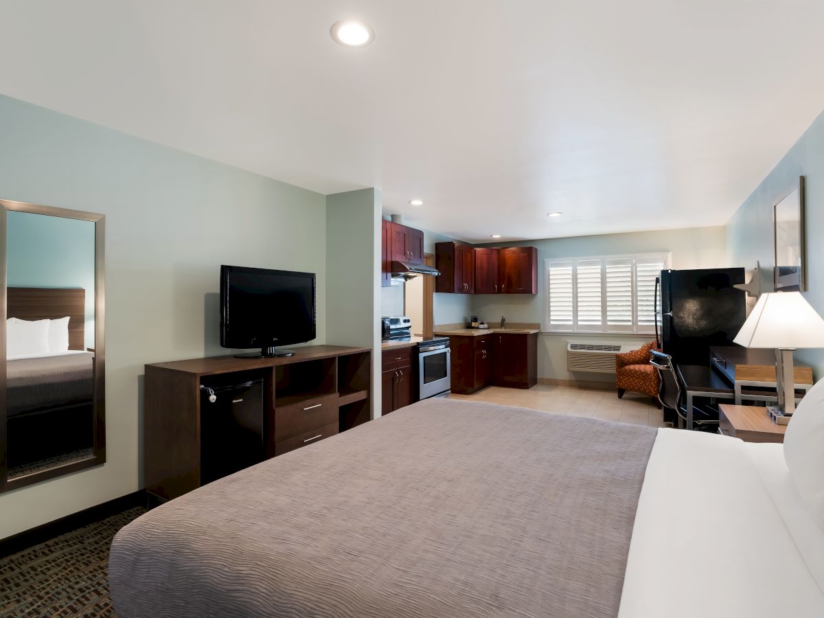 A modern hotel room with a bed, TV, mirror, desk, chairs, lamp, and a kitchenette area with a stove, microwave, and cabinets in the background.