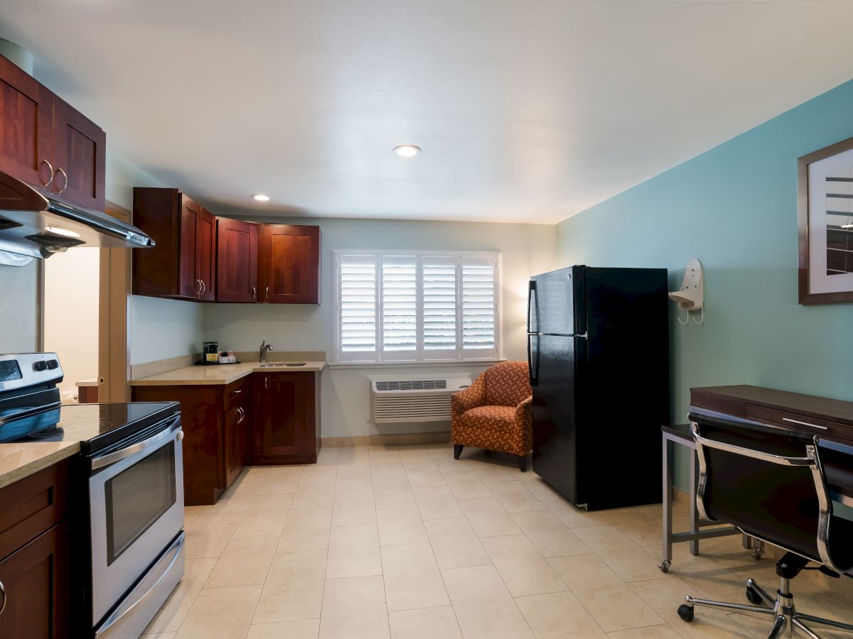 This image shows a modern kitchen and dining area with an oven, refrigerator, desk, chair, and cabinets. The room has a tiled floor and a window.