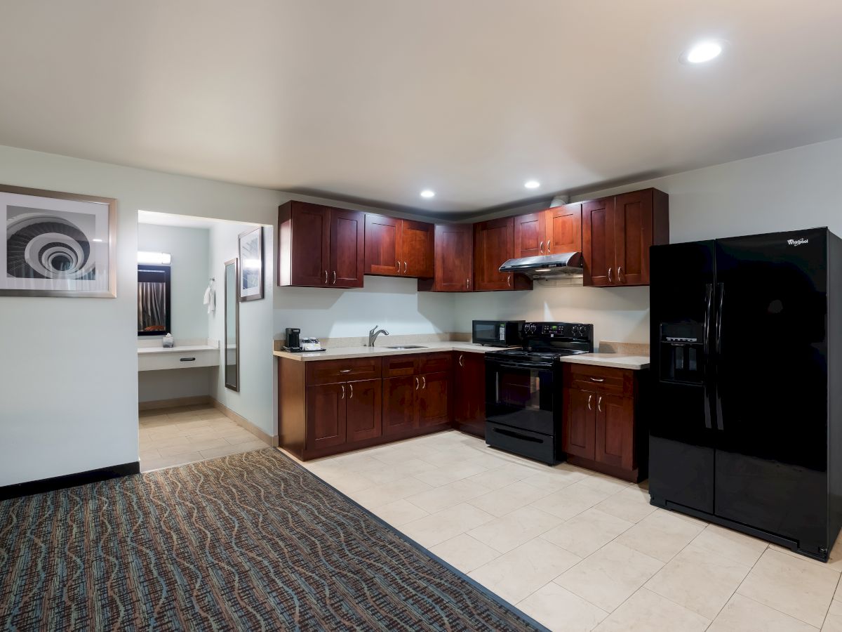 This image shows a modern kitchen with wooden cabinets, a black stove, refrigerator, microwave, and a small sink. It features tiled and carpeted areas.