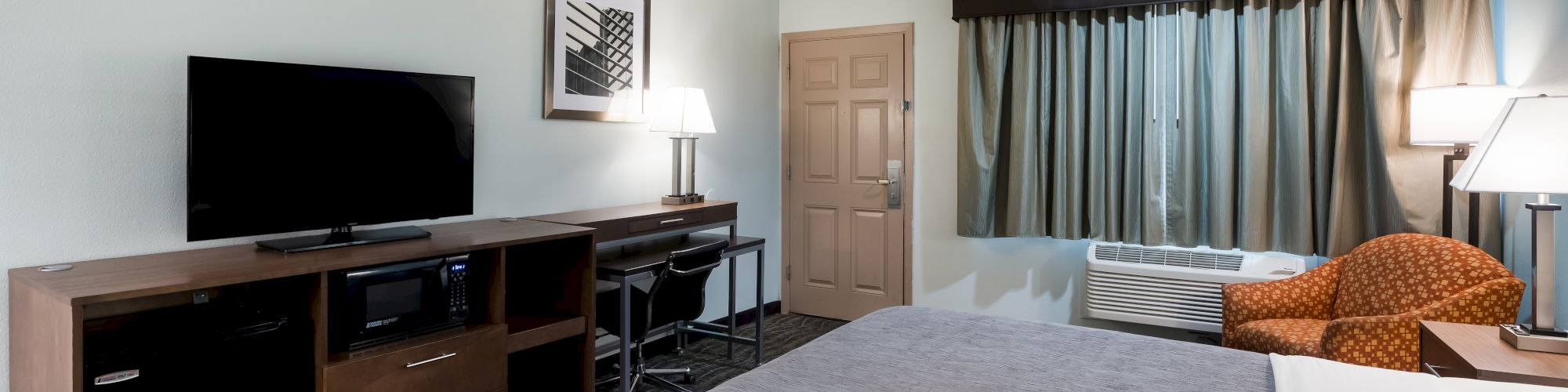 The image shows a hotel room with a bed, a TV on a cabinet, a desk with a lamp, a window with curtains, and an armchair.