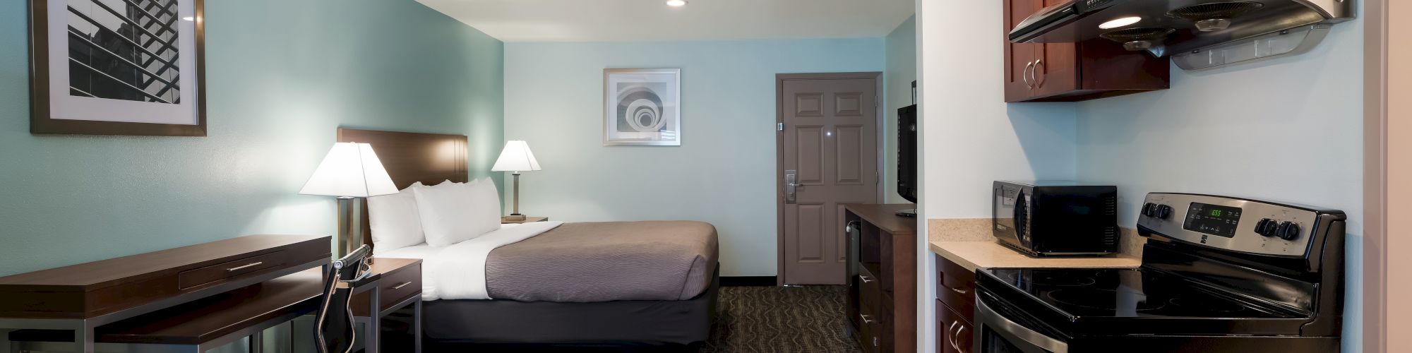 The image shows a hotel room with a bed, desk, and kitchen area, featuring a stove, microwave, and refrigerator, decorated in modern style.