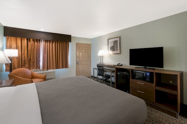 A neatly arranged hotel room with a large bed, a TV on a wooden stand, a desk, armchair, and curtains covering the window, lit by lamps.