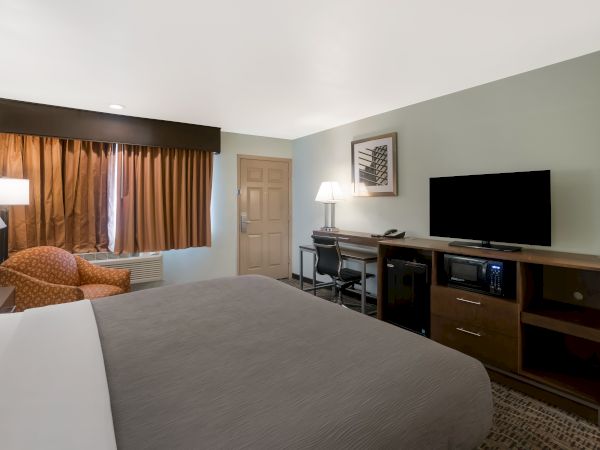 A neatly arranged hotel room with a large bed, a TV on a wooden stand, a desk, armchair, and curtains covering the window, lit by lamps.