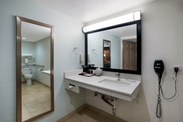 The image shows a bathroom with a sink, large mirror, wall-mounted hairdryer, towels, toiletries, and a toilet visible in the background.