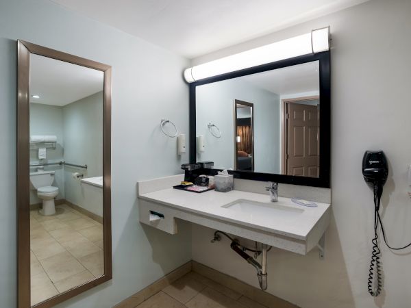 The image shows a bathroom with a sink, large mirror, wall-mounted hairdryer, towels, toiletries, and a toilet visible in the background.