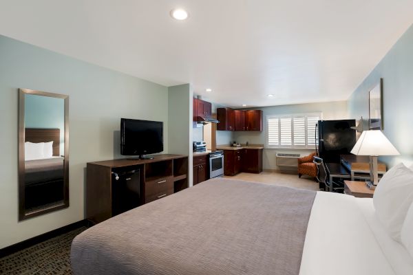 This image shows a hotel room with a bed, TV, dresser, mirror, kitchenette, and seating area. It has modern furniture and lighting in a cozy setup.