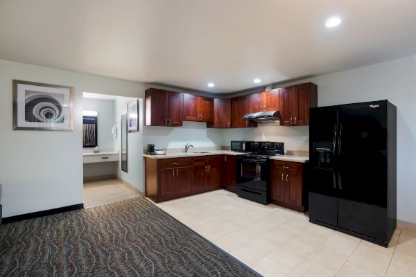 A modern kitchen with dark wood cabinets, black appliances, and adjacent bathroom. The room has tiled and carpeted sections, with framed art on the wall.