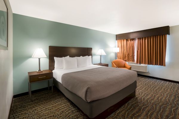 The image shows a hotel room with a large bed, two bedside tables with lamps, an orange armchair, and brown curtains covering the window.