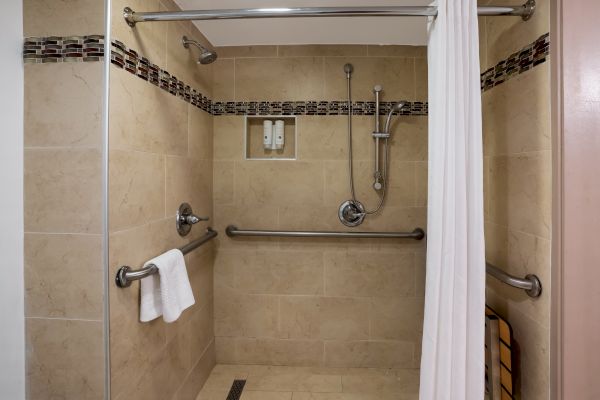 A beige tiled shower with a towel, grab bars, showerhead, and soap dispensers, enclosed by a curtain and glass door ends the sentence.