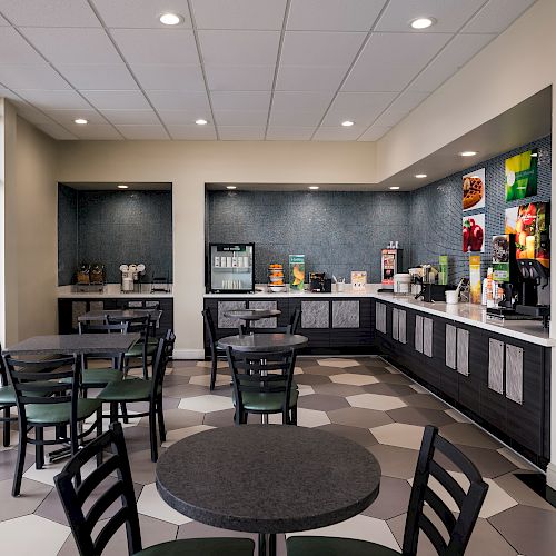 The image shows a small dining area with multiple round tables and chairs, a counter with various beverages and snacks, and large windows bringing in natural light.