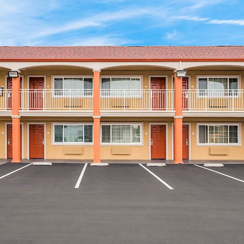 An image of a two-story motel with orange doors, a staircase, balconies, and an empty parking lot under a blue sky.