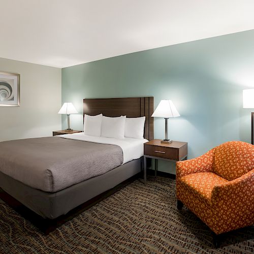 This image shows a hotel room with a neatly made double bed, nightstands with lamps, a wall painting, a patterned armchair, and carpet flooring.