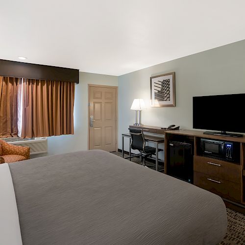 A well-furnished hotel room featuring a bed, an armchair, a desk with a chair, a TV on a cabinet, and a window with curtains.