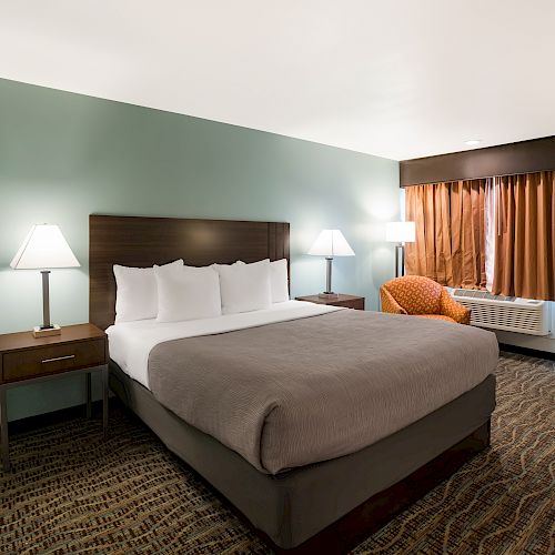 The image shows a neatly made bed in a hotel room with two nightstands, two lamps, a small couch, and a window with curtains.