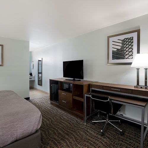 A hotel room with a bed, desk, chair, TV, and lamp. Artwork hangs on the walls, and carpeting covers the floor. The room appears neat and tidy.