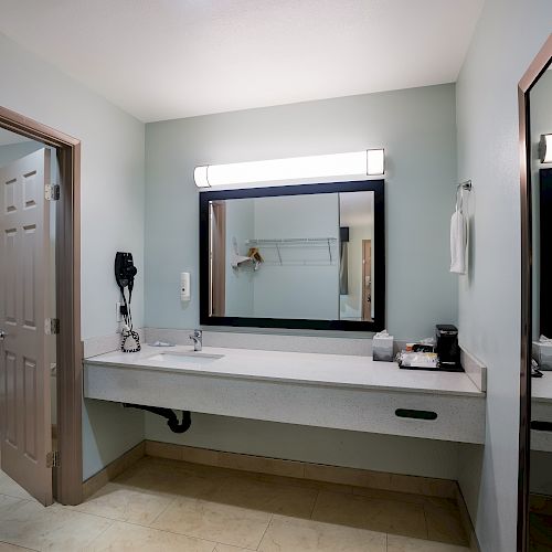 A clean bathroom features a large mirror, countertop with sink, hairdryer, towel rack, and a door leading to a toilet area with additional towels.