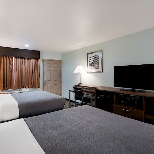 A hotel room with two beds, a nightstand, a TV on a console, a desk with a lamp, and a photo on the wall. The window has brown curtains.