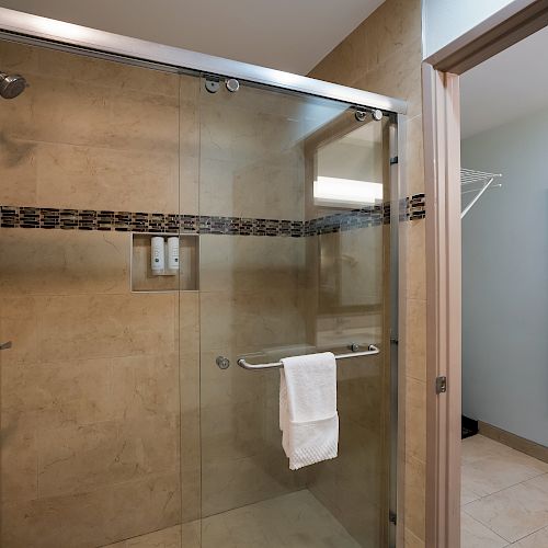 The image shows a modern bathroom with a glass-enclosed shower, tiled walls, and a towel hanging on the door handle. A dressing area is adjacent.