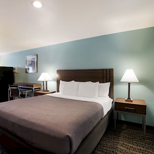 A neatly arranged hotel room with a large bed, bedside tables with lamps, a desk, an armchair, a mini-fridge, and framed artwork on the walls.