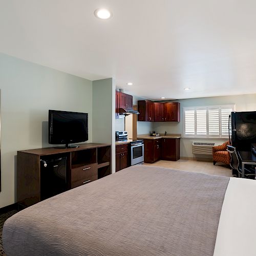 A hotel room with a bed, television, dresser, mirror, kitchenette, desk, chairs, and a lamp, all under bright lighting.