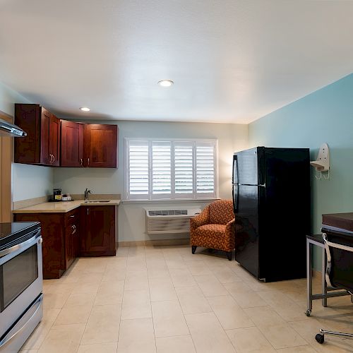 A small kitchen and living area with wooden cabinets, stove, sink, refrigerator, armchair, desk, and chair, with a window and framed picture on the wall.