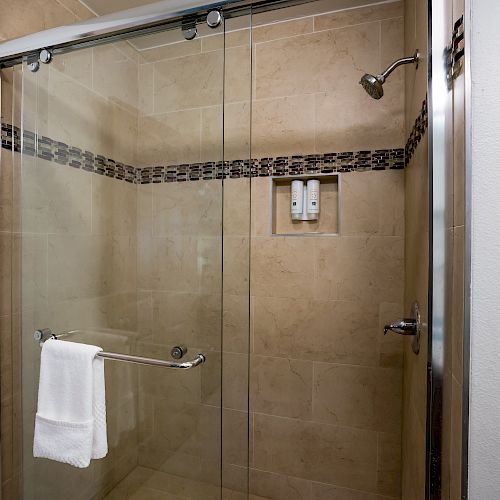 A modern glass-door shower with beige tiles, a towel hanging on a bar, wall-mounted shampoo and soap dispensers, and a showerhead.