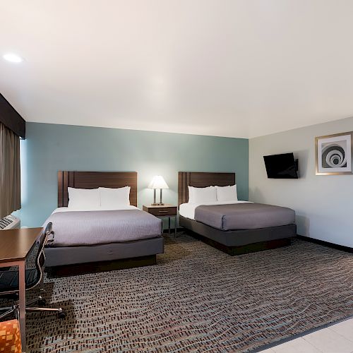 The image shows a hotel room with two double beds, a desk, a chair, a TV, lamps, and a picture frame. The bathroom area is also visible in the background.