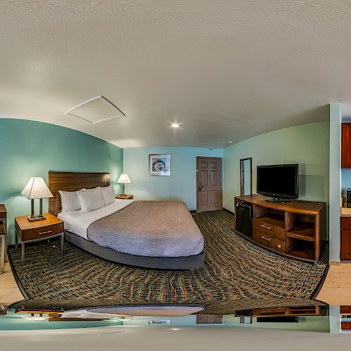 This image shows a modern hotel room with a king-sized bed, a desk, a flat-screen TV, a full kitchen with appliances, and a seating area by the window.