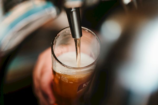 A hand holds a glass as beer is being poured from a tap, forming a foamy head. The scene appears to be taken at a bar or pub.