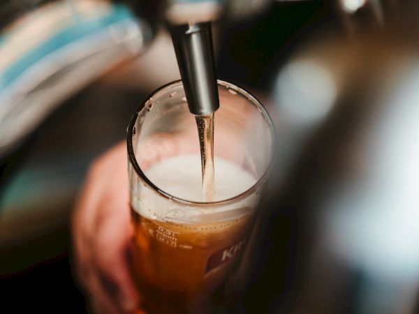 A hand holds a glass as beer is being poured from a tap, forming a foamy head. The scene appears to be taken at a bar or pub.