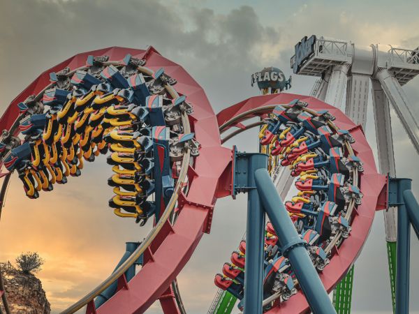 The image depicts a roller coaster with loops and another ride in the background, creating a thrilling amusement park setting at sunset.